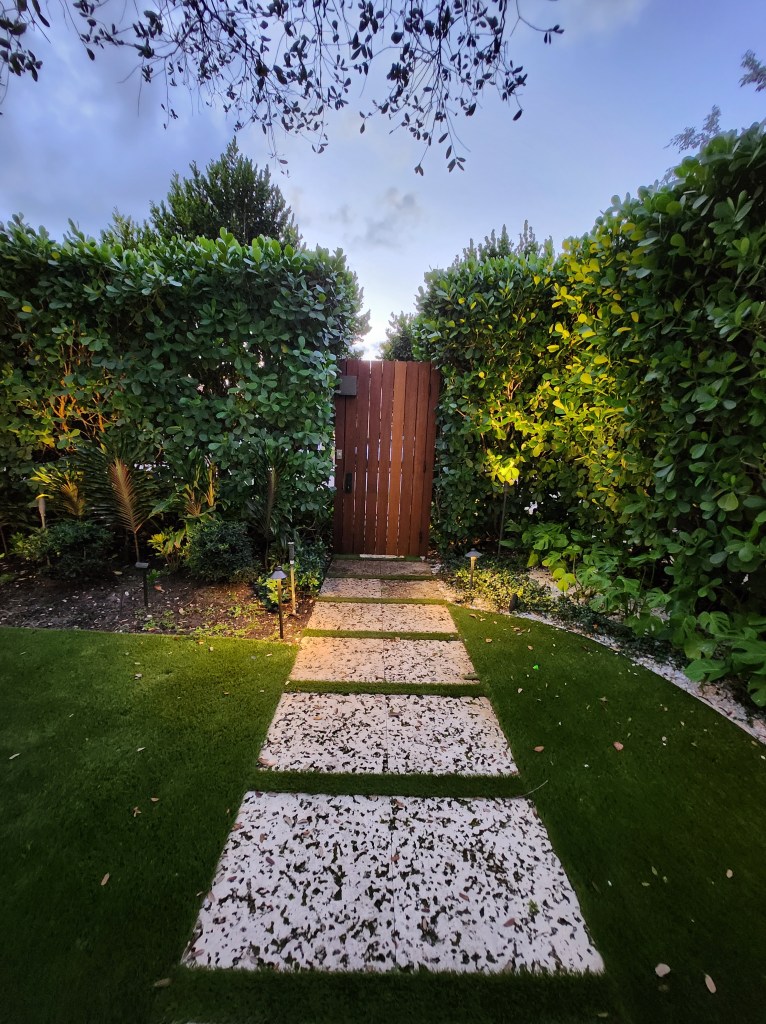 A serene garden path leading to a wooden gate, framed by lush green hedges and soft lighting.